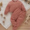 A soft, bick red cable knit loungewear set for babies and toddlers, including a long-sleeved jumper and matching joggers, laid flat on a white blanket with wooden toys.