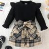 A smart two-piece girls' outfit, featuring a long-sleeved black ribbed top with ruffle shoulders, and a matching beige and black tartan skirt with a large bow.