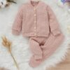 A soft, pink-coloured two-piece cable knit baby outfit, including a button-front cardigan and matching cuffed trousers, laid flat on a fluffy white rug.