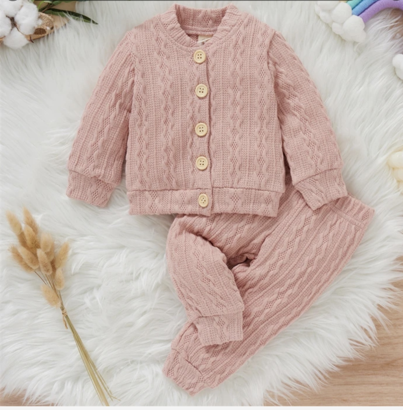 A soft, pink-coloured two-piece cable knit baby outfit, including a button-front cardigan and matching cuffed trousers, laid flat on a fluffy white rug.
