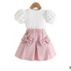 A pretty two-piece girls' outfit, featuring a white ribbed top with quilted puff sleeves and a pink pleated skirt with two large bows and an elasticated waist.