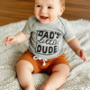 A baby and toddler boy's two-piece set, featuring a grey marl cotton t-shirt with "Dad's Little Dude" in black print and a pair of rich tan shorts with a drawstring.